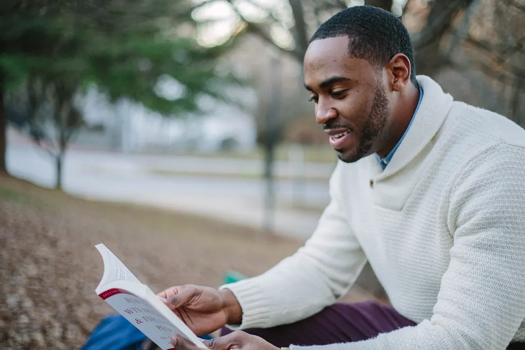 Homme souriant, plongé dans un livre, assis dehors en pull blanc