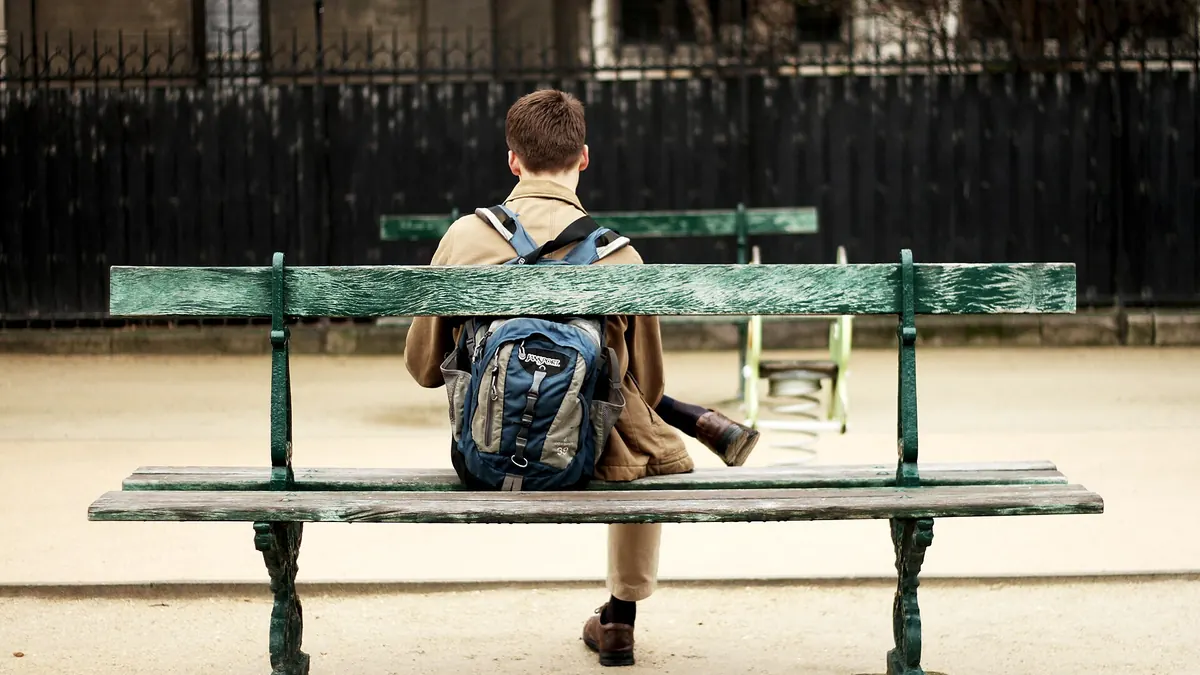 Jeune homme assis seul sur un banc de parc, vu de dos, sac sur l'épaule