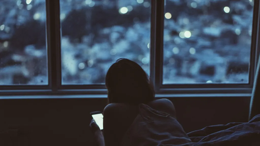 Femme vue de dos devant une fenêtre, le soir, son téléphone à la main